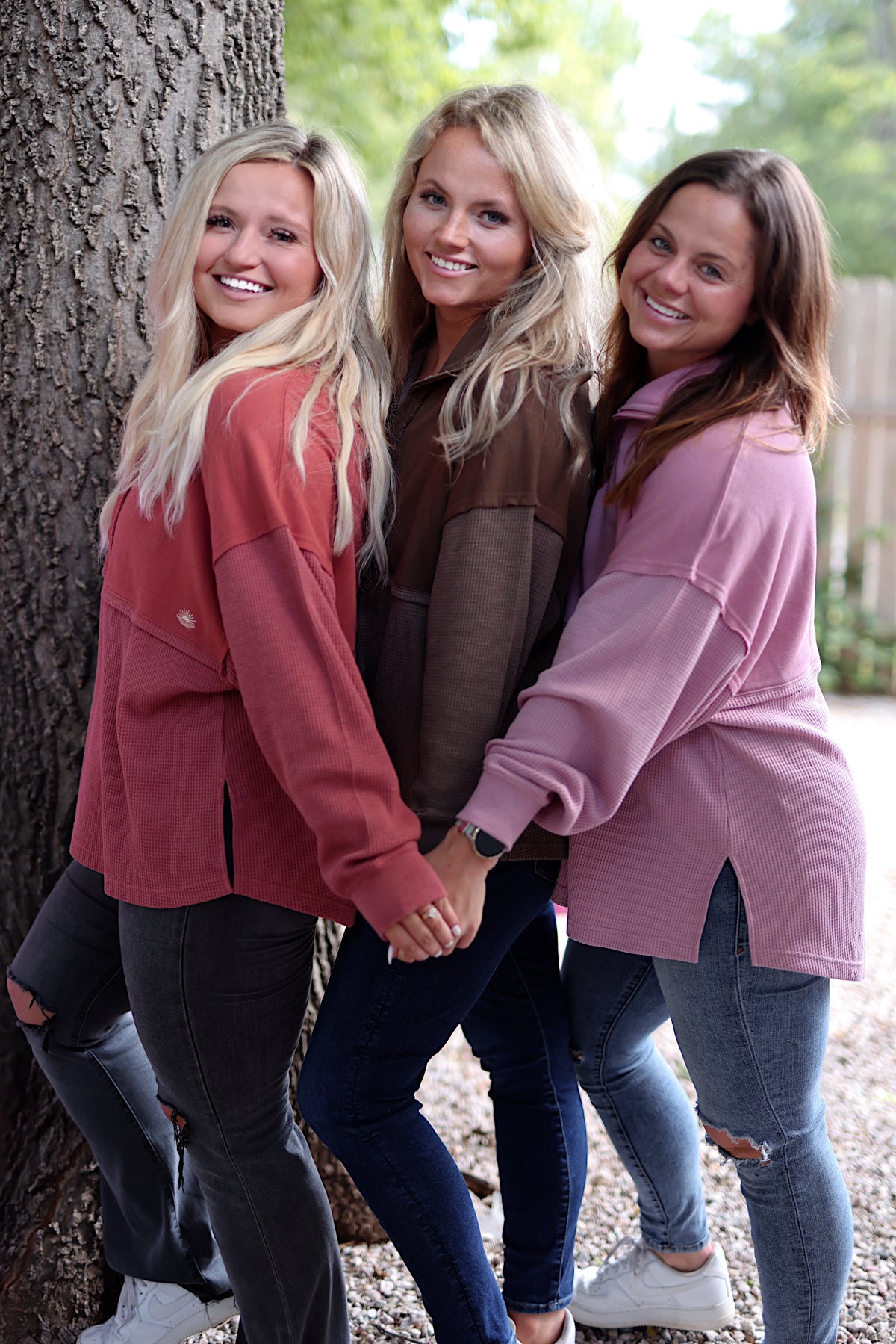 Three women wearing cozy oversized sweaters in pink, rust, and brown, styled with jeans against a tree.