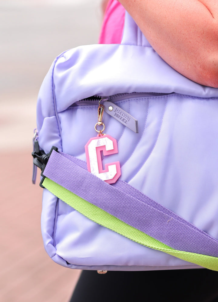 A person carrying a large lavender quilted duffel bag with a neon green strap and a pink initial 'C' charm, set against a blurred outdoor background.
