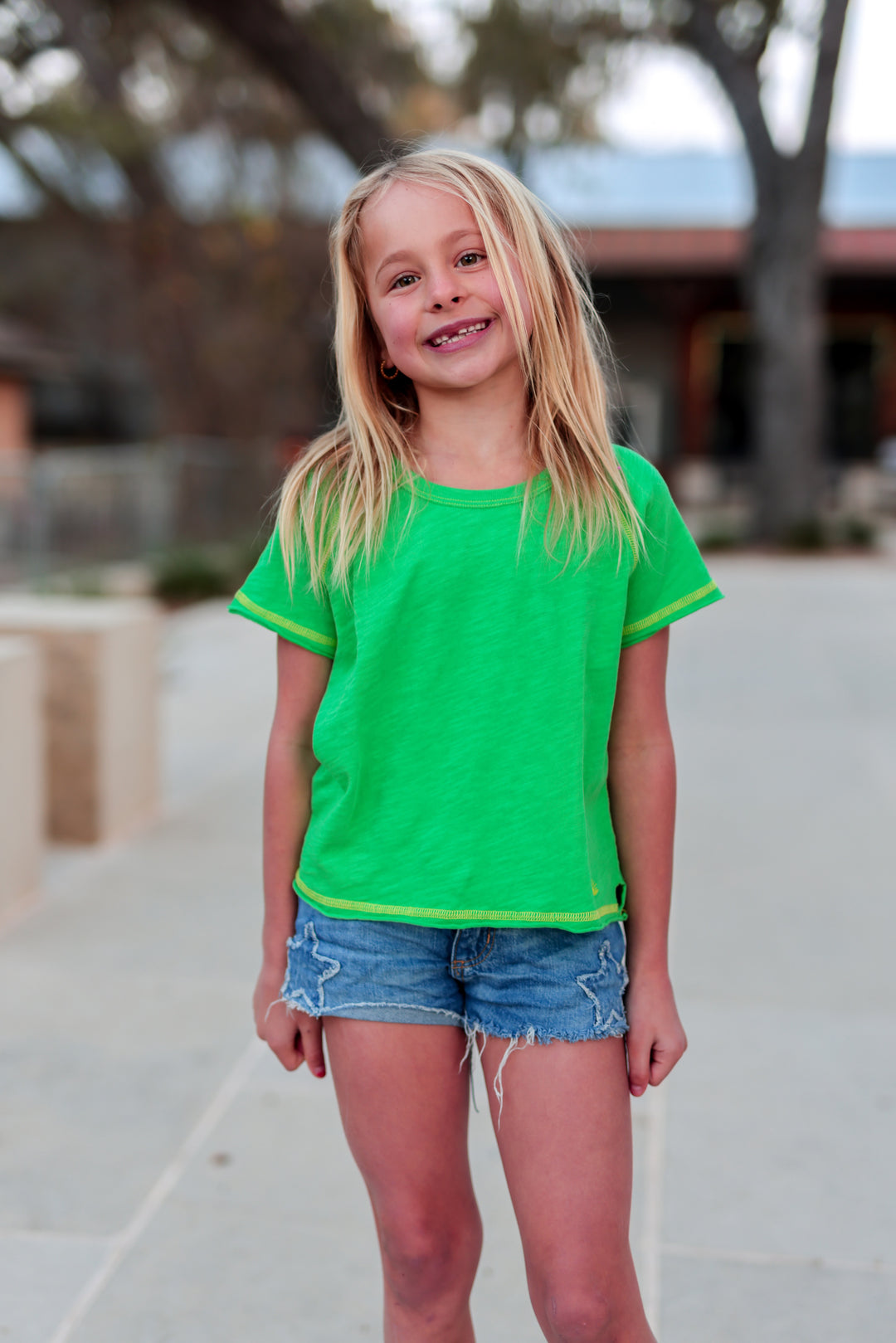Young girl wearing a green shirt and denim shorts standing outdoors.