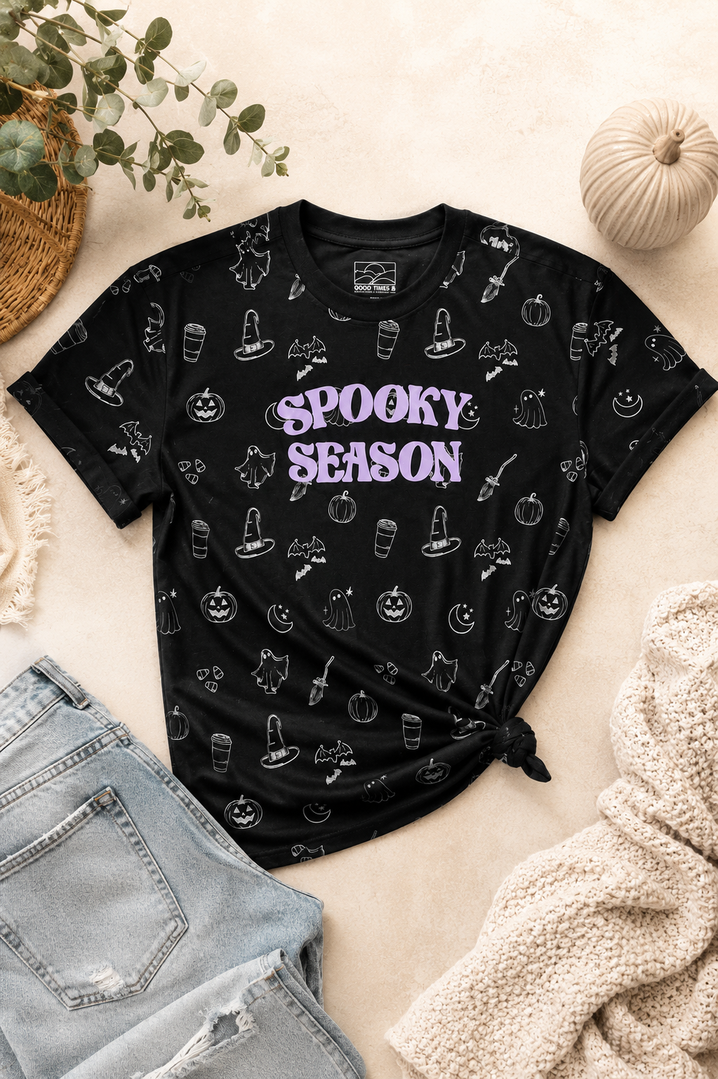A black short-sleeve graphic T-shirt laid flat on a beige surface, surrounded by seasonal props including a small pumpkin, eucalyptus leaves, denim shorts, and a cream knit blanket. The shirt has an allover white spooky line-art print and large purple text reading “SPOOKY Season” across the chest.