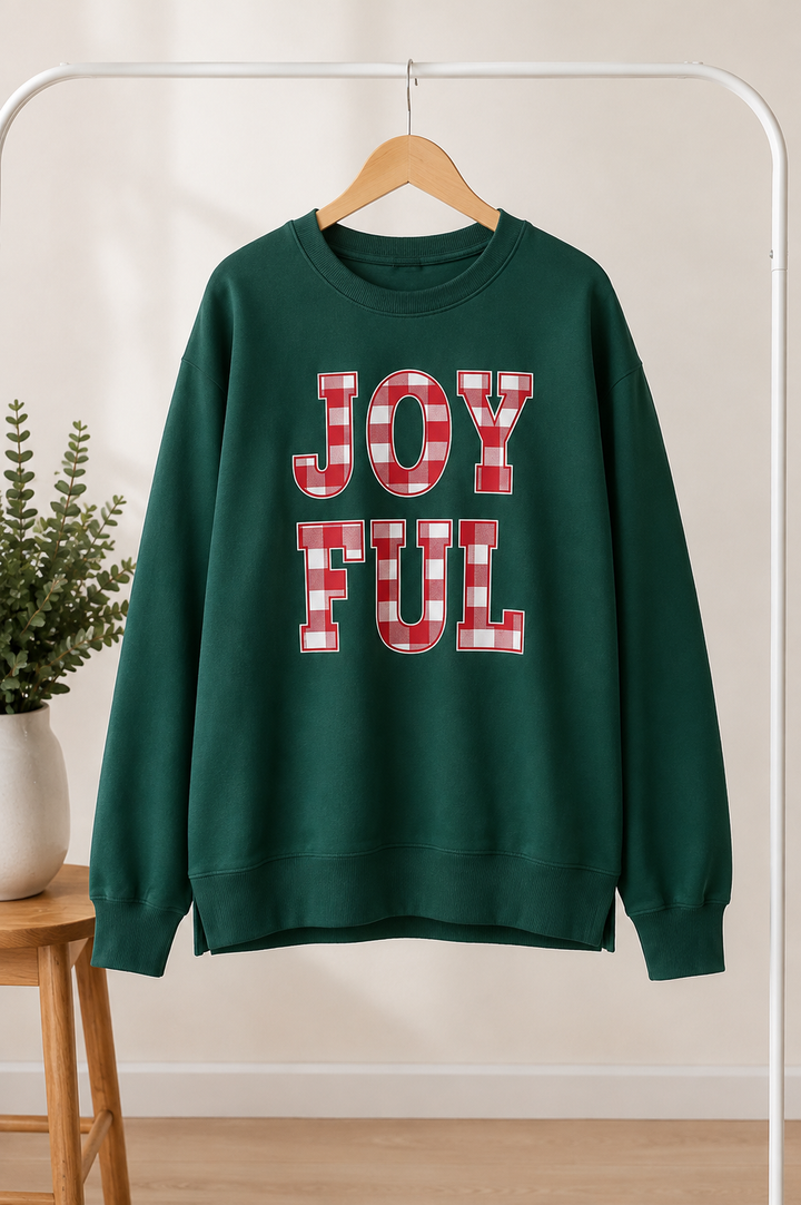 A dark green crewneck sweatshirt hanging on a wooden hanger from a white clothing rack. The sweatshirt features the words 'JOYFUL' printed in large block letters with a red and white buffalo plaid pattern.