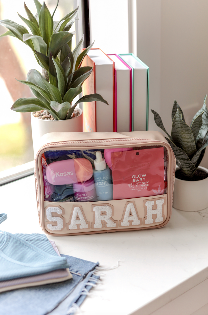 Blush pink cosmetic bag with 'SARAH' label, containing skincare products and plants in the background.