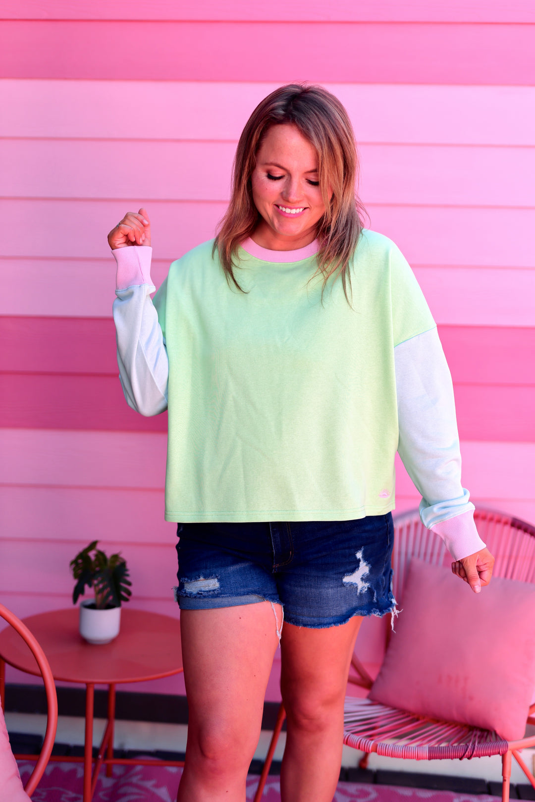 Woman wearing a light green sweater and denim shorts standing in front of a pink wall.