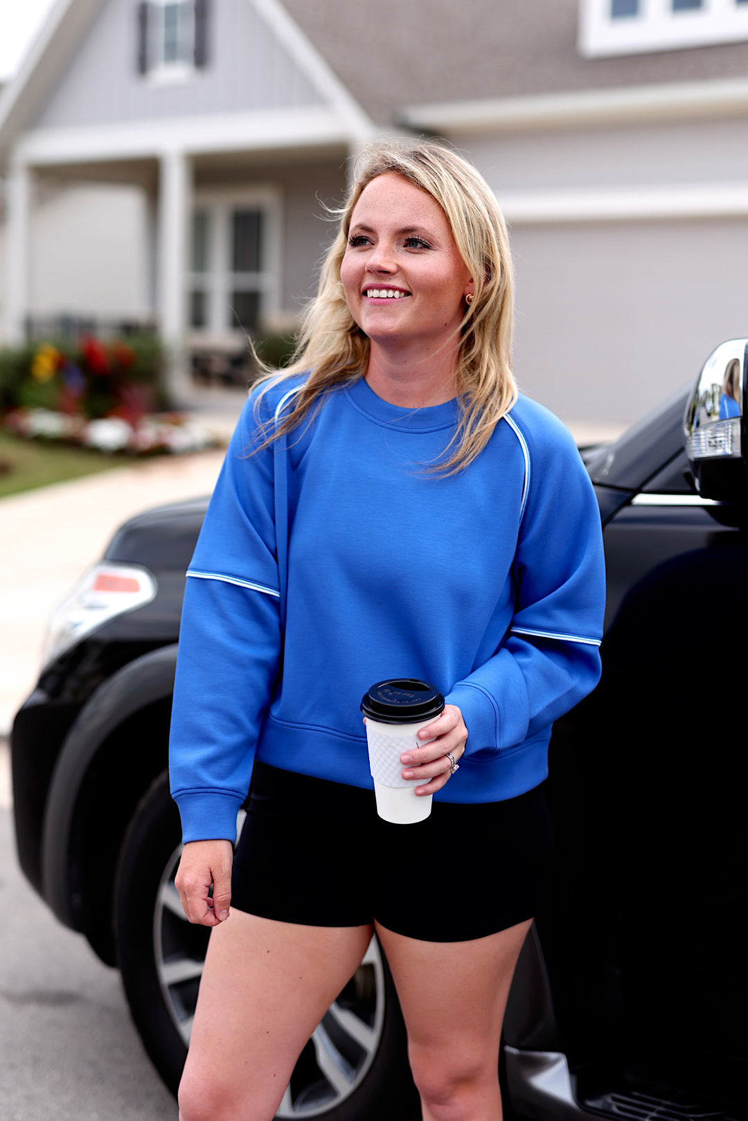 Woman in a blue sweatshirt and black shorts, holding coffee outside a modern home.