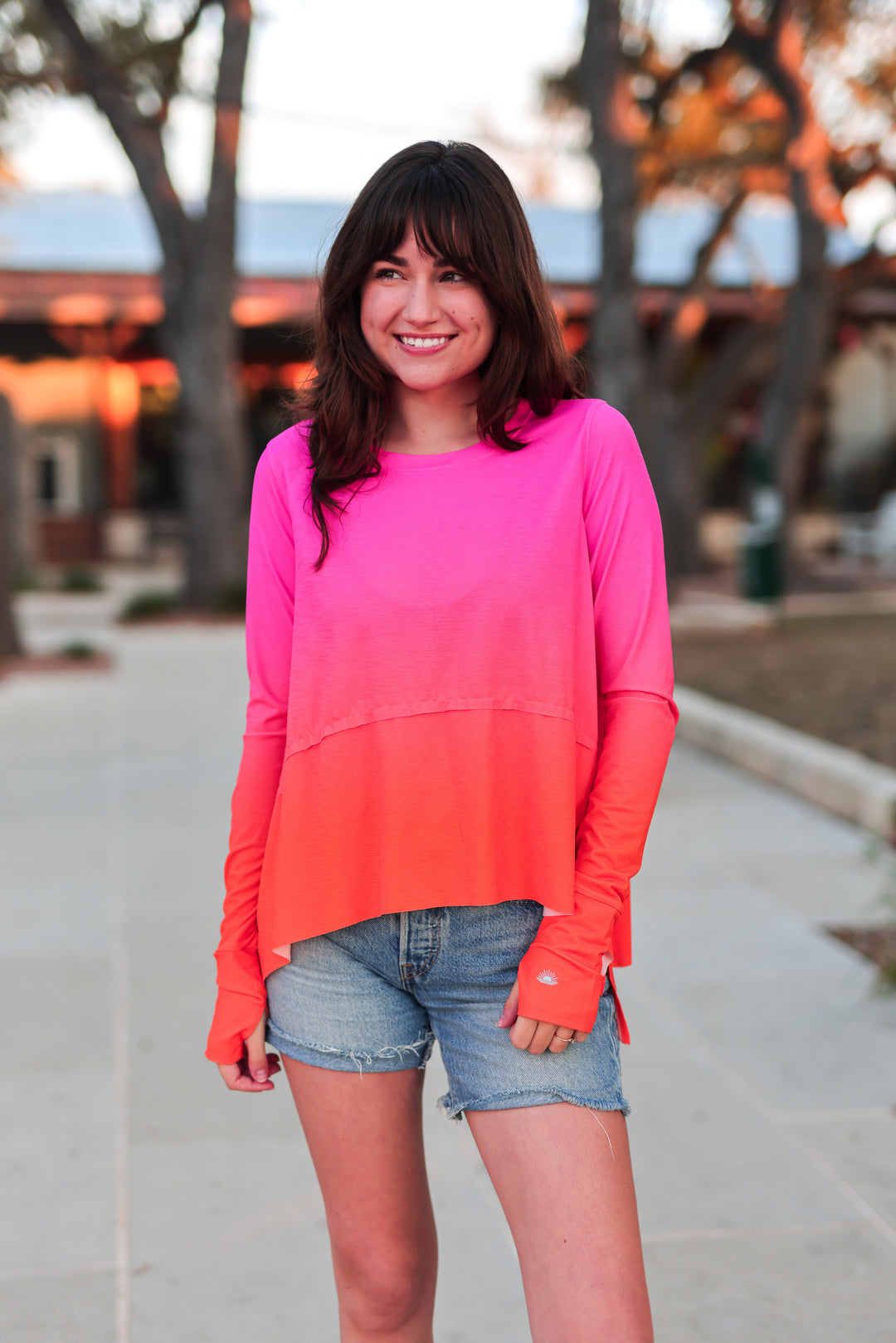 Woman wearing a pink and orange gradient long sleeve top with denim shorts outdoors.