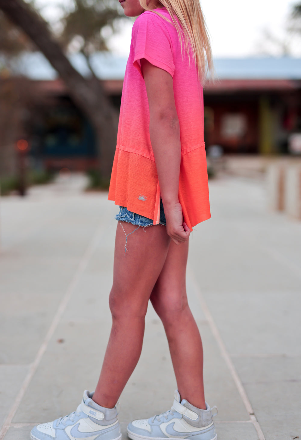 Girls' short sleeve ombre t-shirt in pink and orange, casual style, paired with denim shorts and sneakers.
