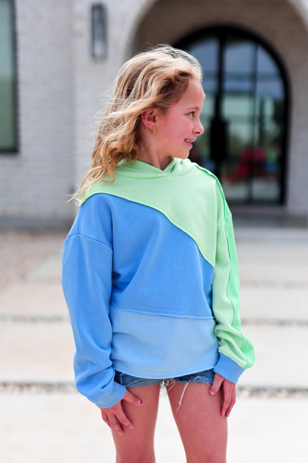 Young girl wearing a blue and green color-blocked hoodie in front of a building.
