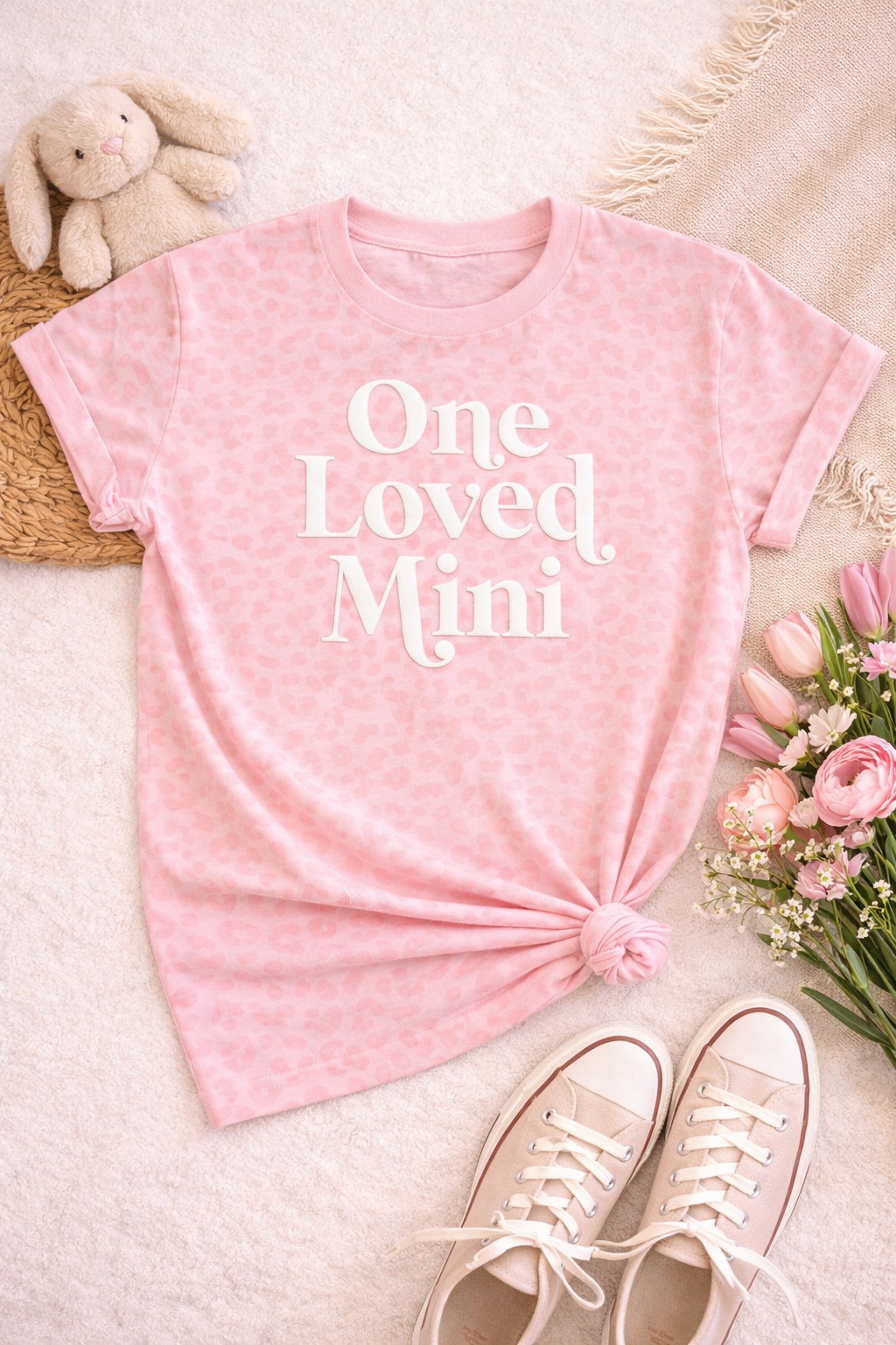 Pink t-shirt with 'One Loved Mimi' text on a light surface with shoes and flowers.