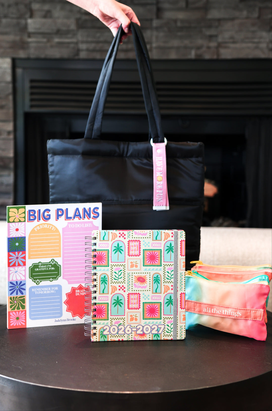 Black tote bag with pink keychain, colorful planner, and pouch on a table against a stone wall background.