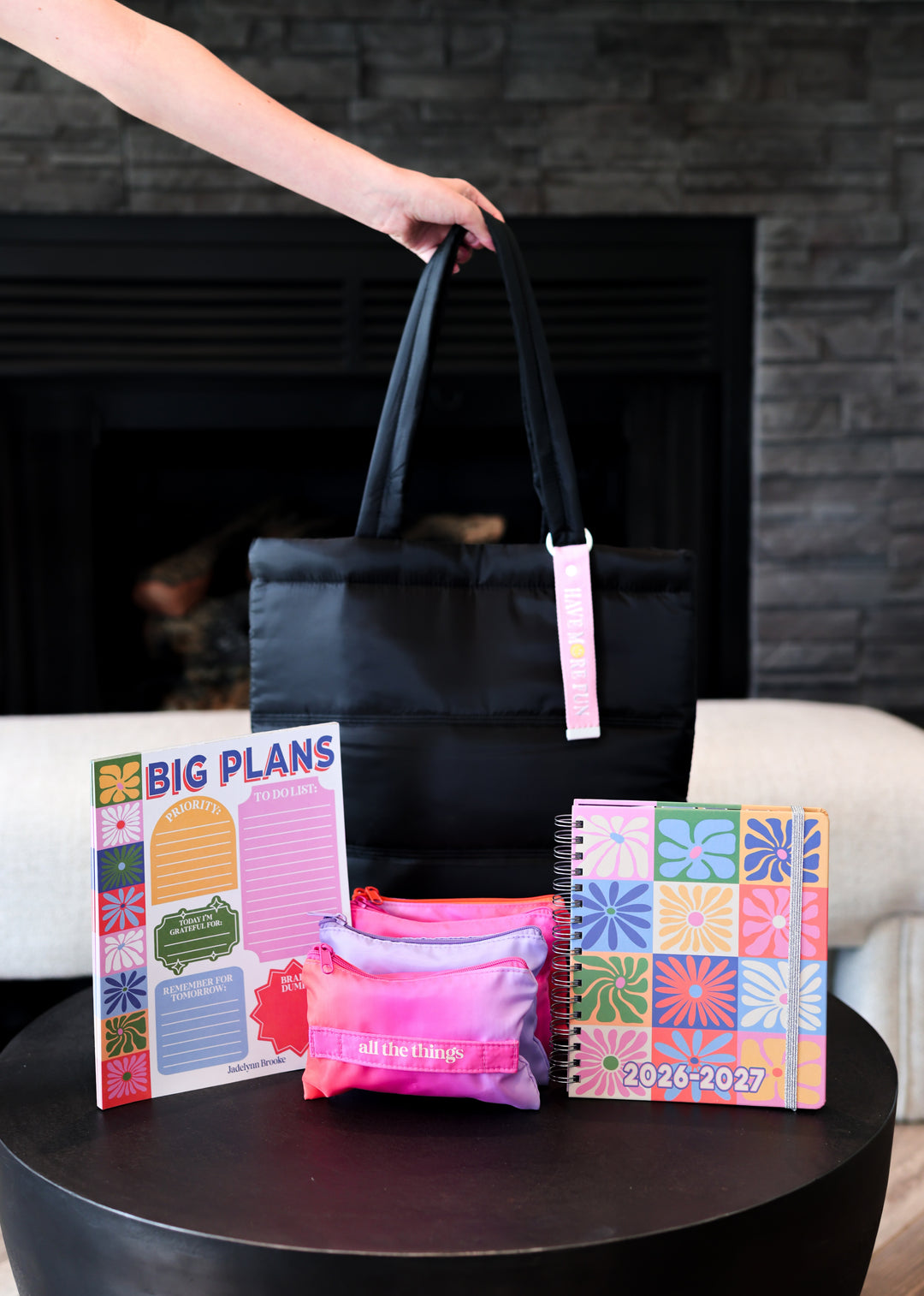 Black tote bag with pink accents on a table next to planners and a notebook.