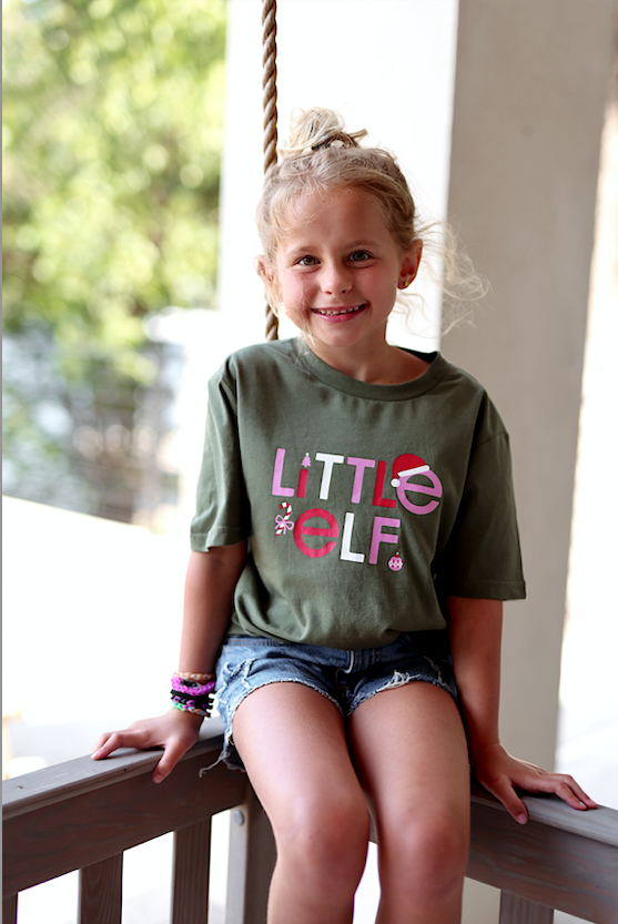 Child wearing an olive green Little Elf graphic t-shirt, paired with denim shorts, sitting on a swing.