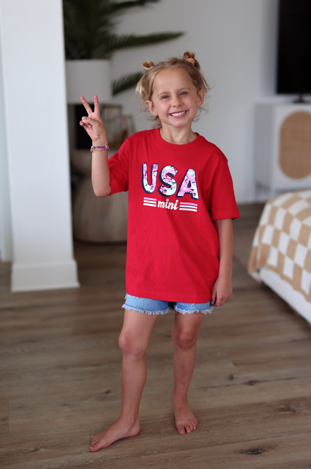 Child in a red USA mini t-shirt with floral print, smiling and making a peace sign, paired with denim shorts.