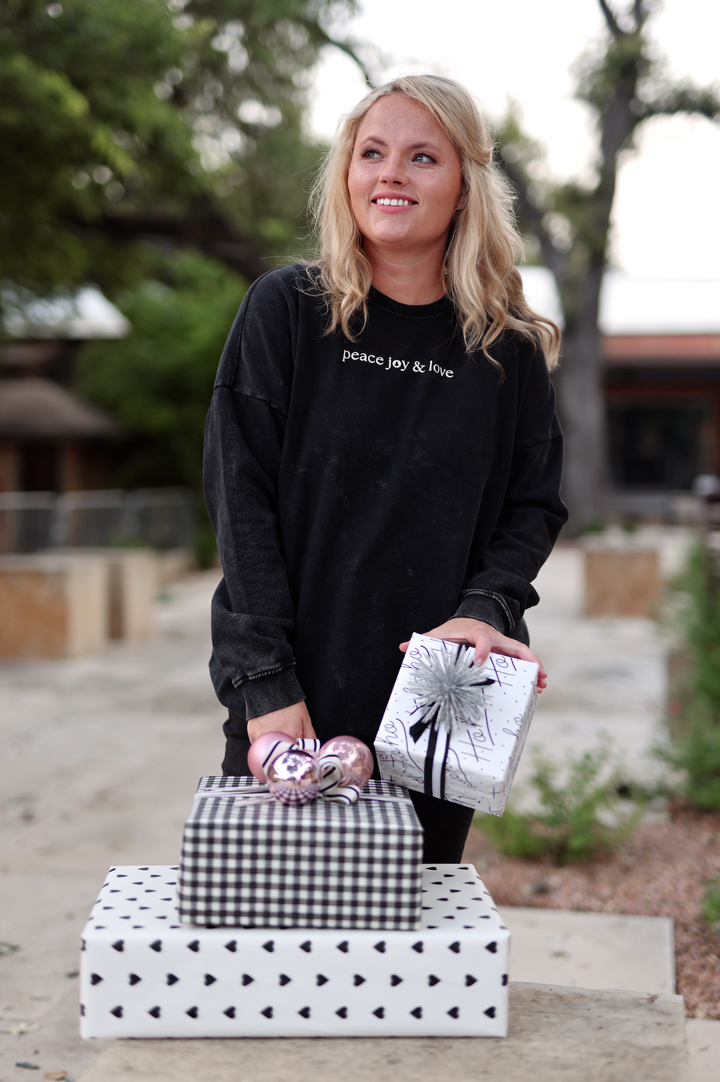 Model wearing black oversized sweatshirt 'peace joy & love', holding decorative gift boxes and ornaments.