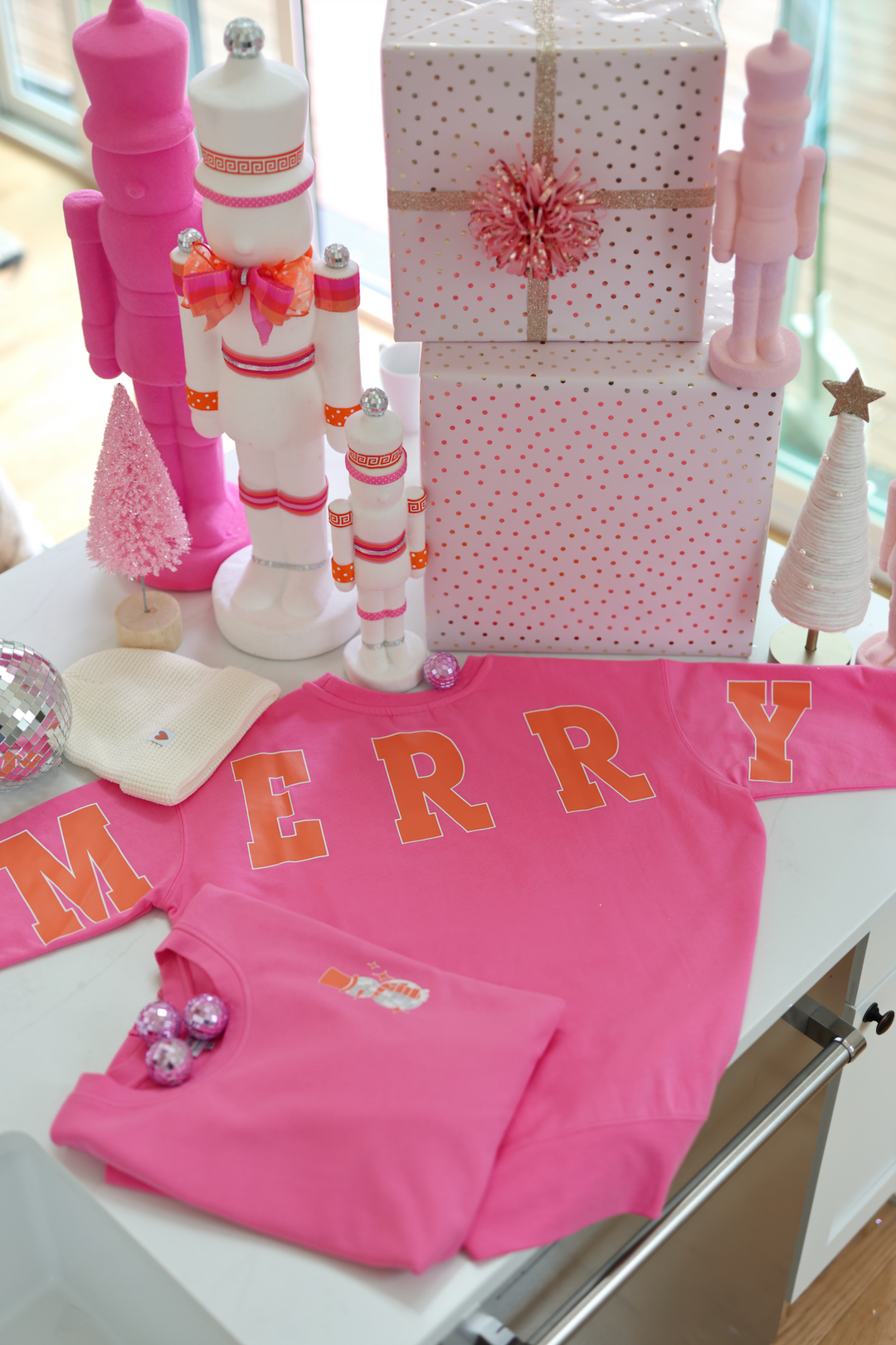 Pink sweatshirt with 'MERRY' print, cozy fabric, styled on a festive table with decorations.