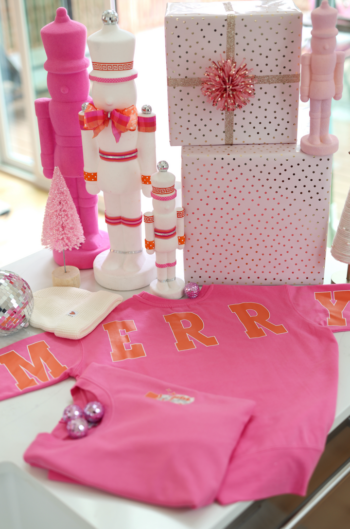Pink sweatshirt with 'MERRY' lettering, surrounded by festive decor and Christmas gifts.