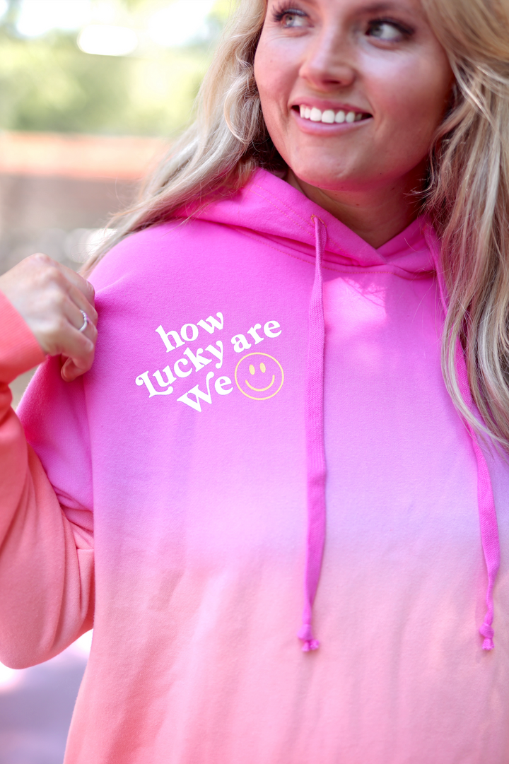 Pink ombre hoodie with 'how lucky are we' text and smiley face, perfect for casual wear.
