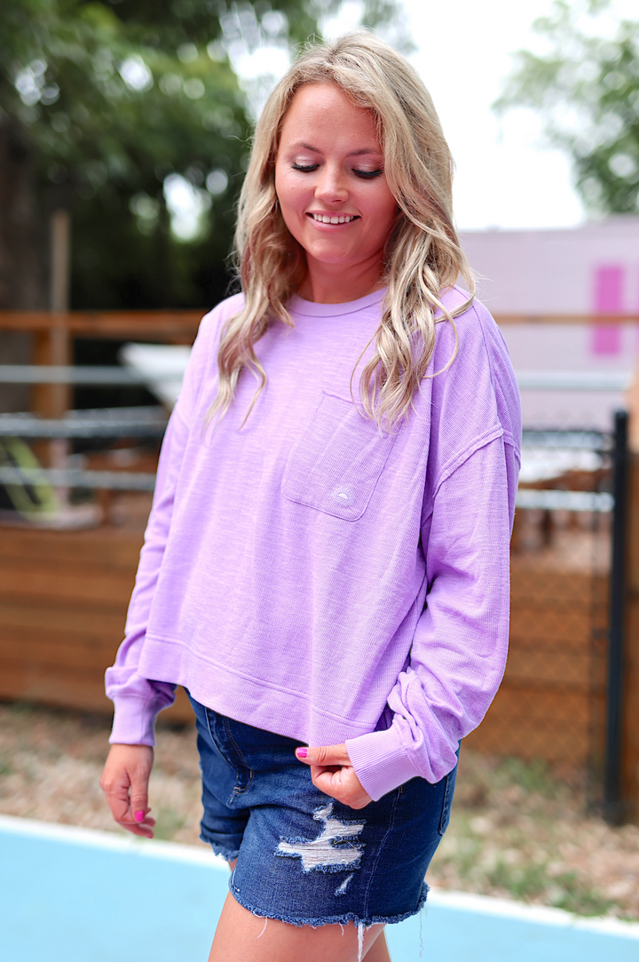 Women's lavender long-sleeve top with pocket, paired with denim shorts for a casual look.
