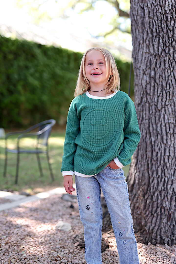 Child in green waffle knit sweatshirt with smiley face design, paired with embroidered light wash jeans.