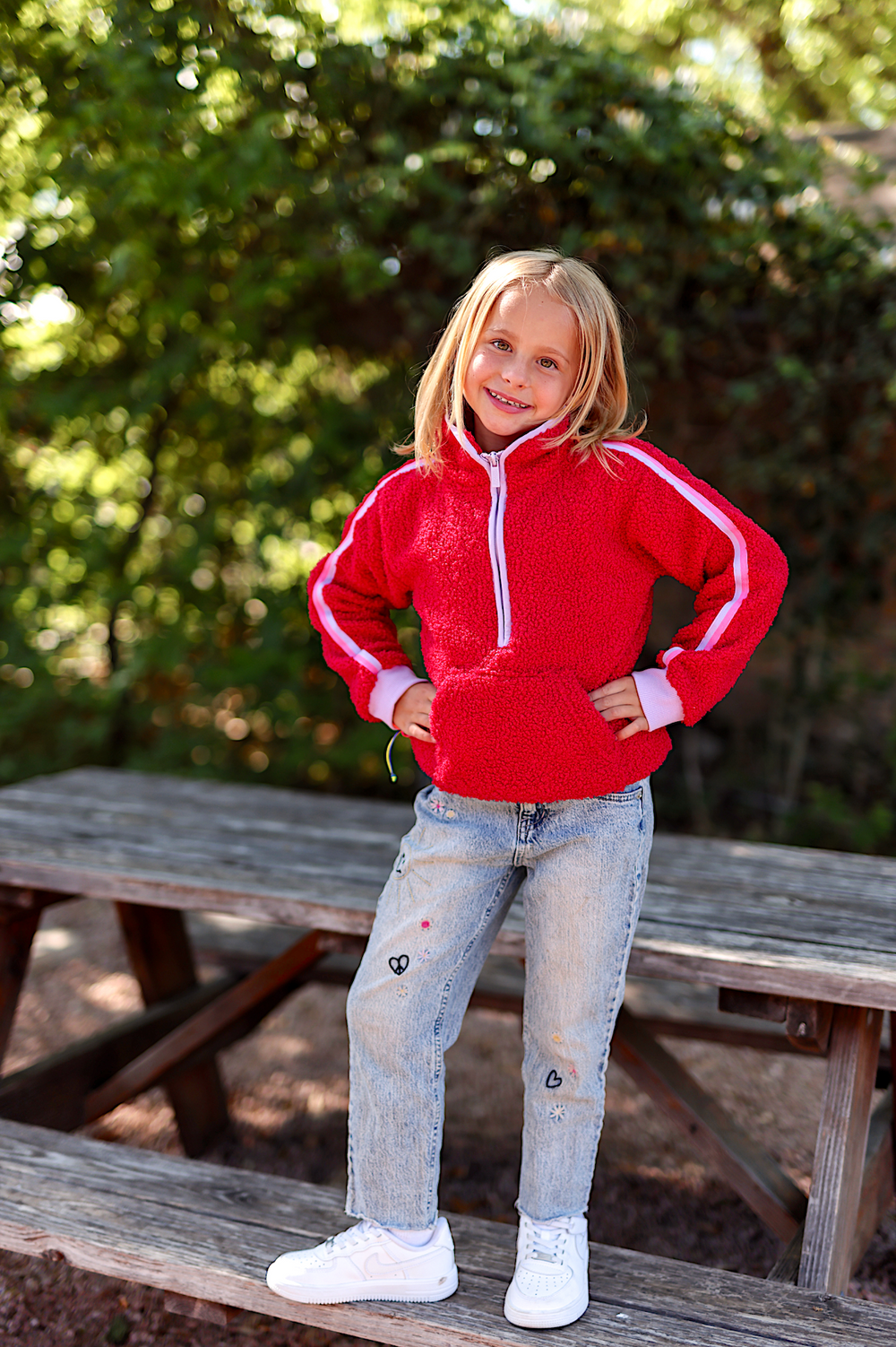 Red fleece zip-up hoodie with white stripes, paired with embroidered jeans and white sneakers on a girl.