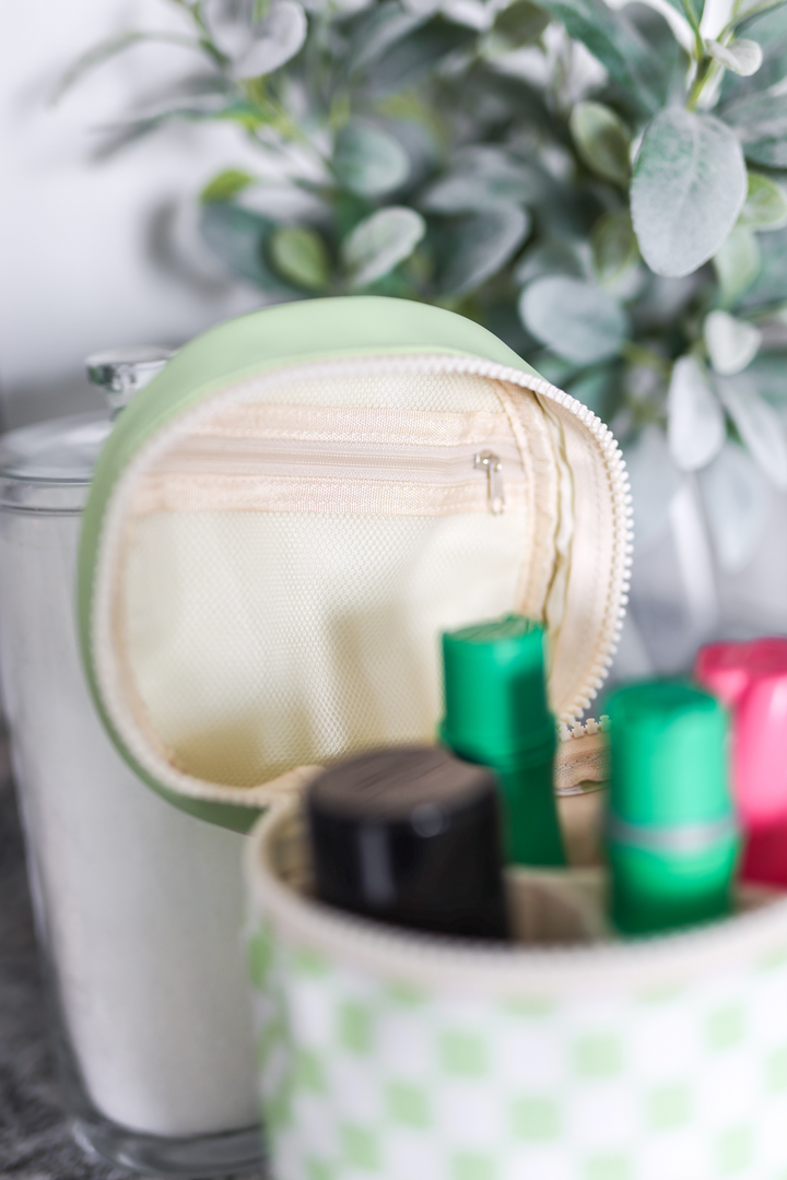 Small round green pouch with a zipper, containing various items including a black container and green boxes, with a blurred plant in the background.