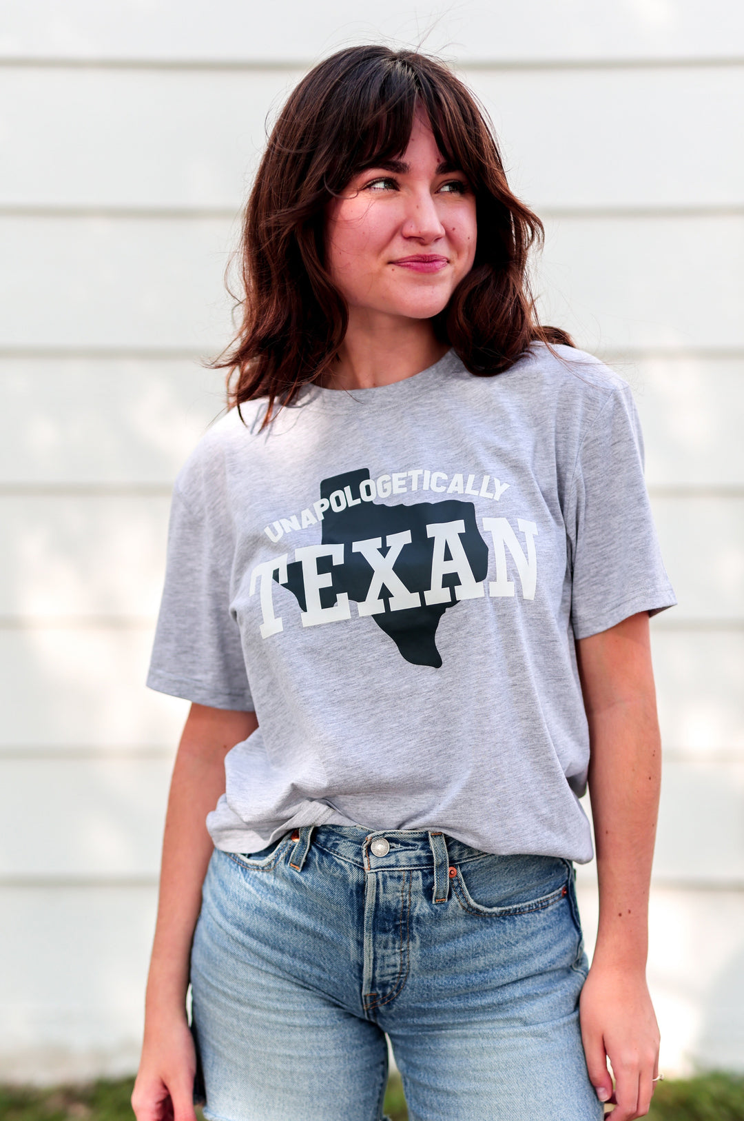 Person wearing a gray t-shirt with 'Unapologetically Texan' text and denim shorts outdoors.