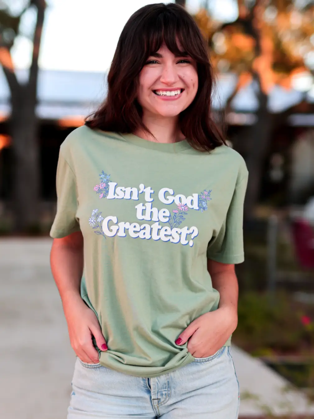 Woman wearing a green t-shirt with 'Isn't God the Greatest?' text and floral designs outdoors.