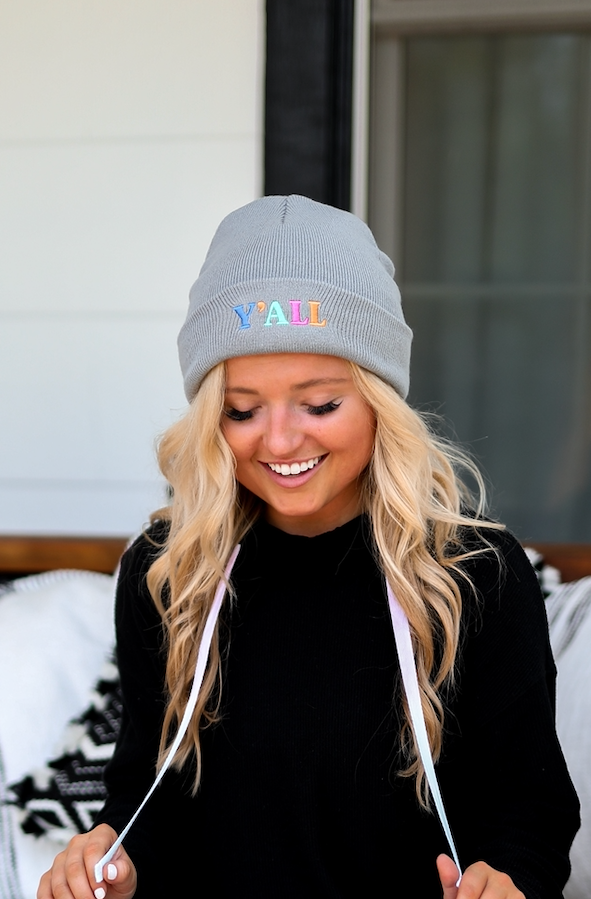 Woman wearing a gray beanie with 'Y'ALL' stitched, black sweater, ripped jeans, seated outdoors on a couch.